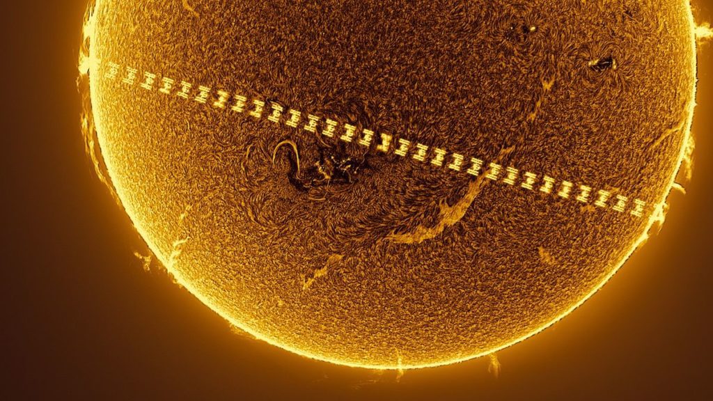 a t-shaped space station passes in front of a fiery sun